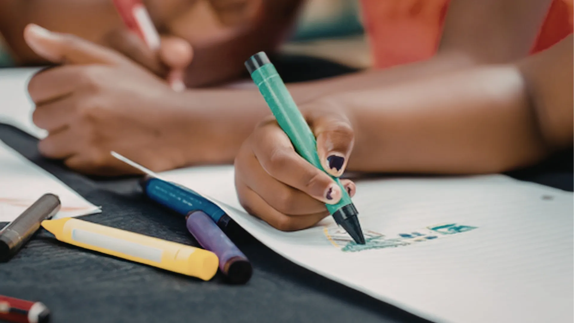 Gros plan sur les mains d'un enfant qui dessine au feutre vert sur un cahier, entouré de feutres de couleur posés sur la table.