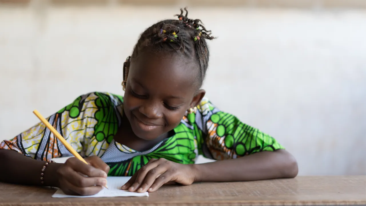 Une fillette souriante, assise à une table en bois, écrit au crayon sur une feuille de papier.
